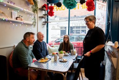 Customers in window seat. Photo of three customers sitting at the window table being served. Food and drink on the table, fairy lights hanging from the makers shelf.