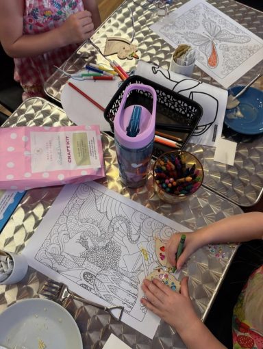 Table of customers colouring in. Customers enjoying a colouring activity with an opened craft kit, drinks and plates on the table.