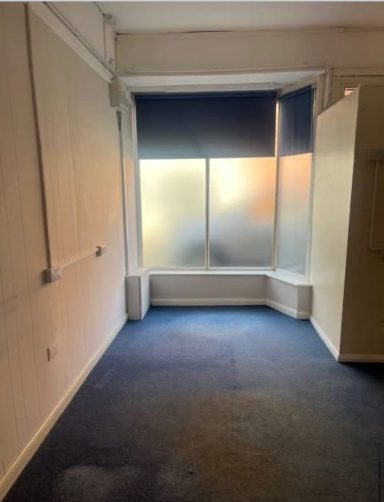 Before: Window bay Before: Window bay with blue blinds half down, off white walls, blue carpet with entry way wall to the right of window bay.