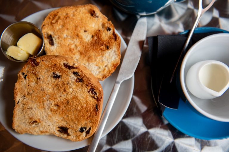 A toasted teacake, open-side up on a plate with a butter knife, small metal pot of butter, empty teacup to the side with a small jug of milk