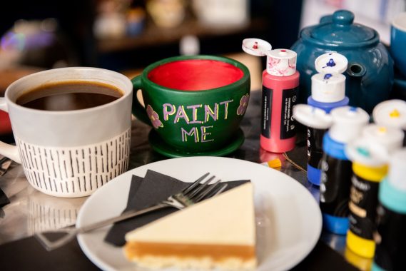 Painting activity with coffee and cake. A dark green ceramic cup with 'Paint Me' written and two flowers. A cup of coffee, teapot, slice of cake and different colour paints.