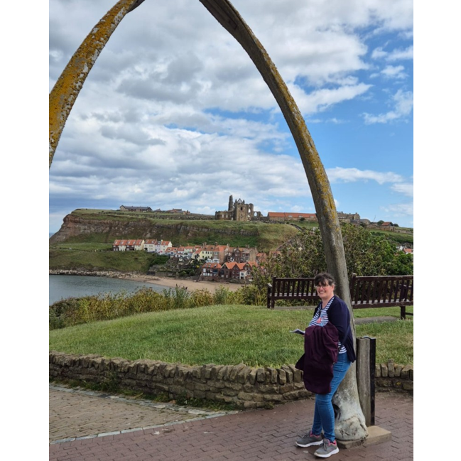 Photo of Jo. Photo of Jo in front of a scenic background with grass, water and a blue sky with many white clouds.