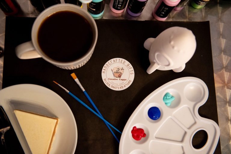 A white rocket ship, baby blue, dark blue and red paint set up with brushes for a customer with a coffee and a slice of cake to the side.