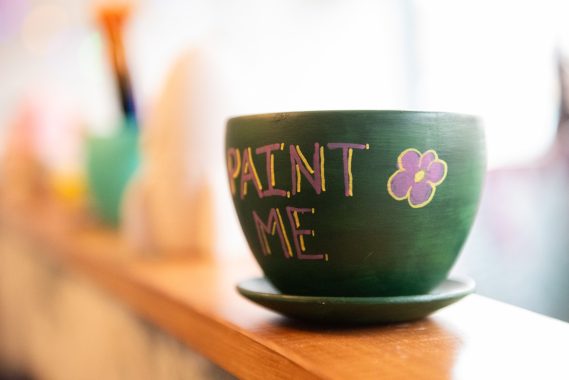 A painted ceramic cup. A ceramic cup painted a dark green with 'Paint Me' written on it and a small flower, blurred background.